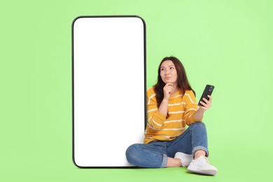 Image of Thoughtful woman with smartphone sitting near big mobile phone with blank screen on light green background. Mockup for design