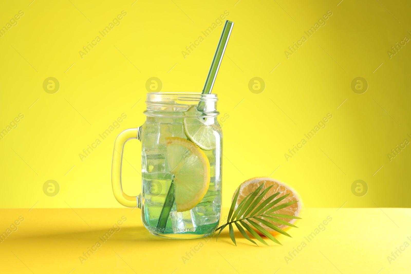 Photo of Tasty refreshing drink with straw in mason jar, citrus fruit and palm leaf on yellow background