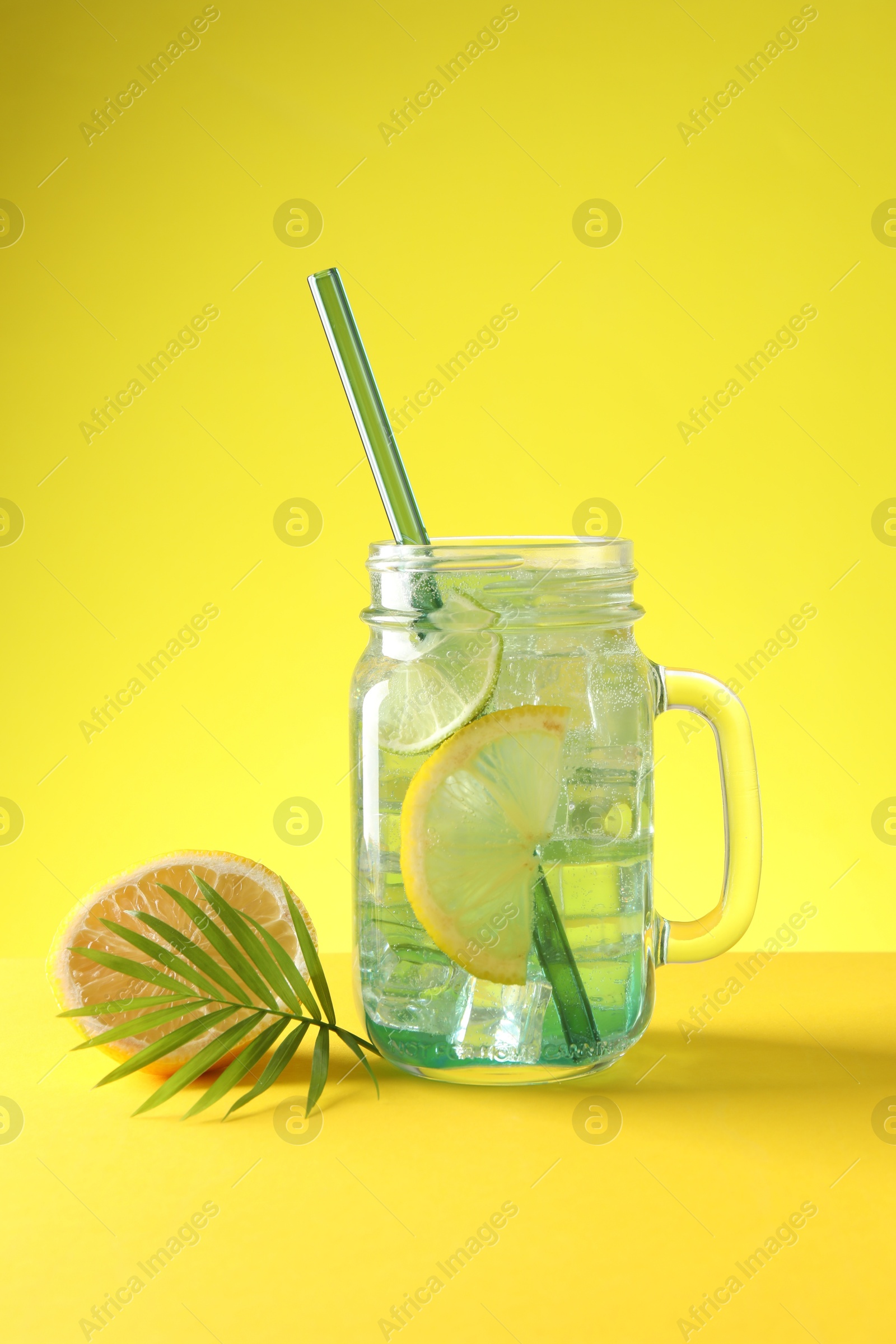 Photo of Tasty refreshing drink with straw in mason jar, citrus fruit and palm leaf on yellow background