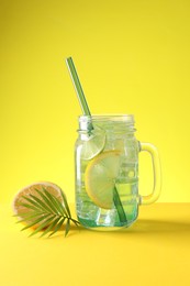 Photo of Tasty refreshing drink with straw in mason jar, citrus fruit and palm leaf on yellow background