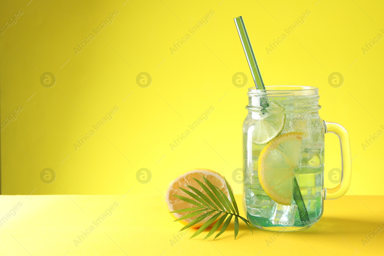 Photo of Tasty refreshing drink with straw in mason jar, citrus fruit and palm leaf on yellow background, space for text