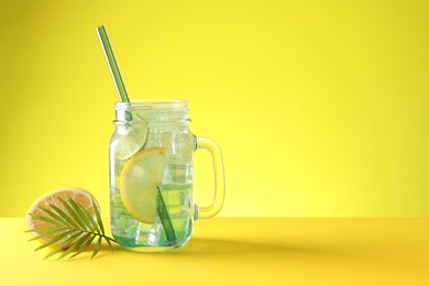 Photo of Tasty refreshing drink with straw in mason jar, citrus fruit and palm leaf on yellow background, space for text