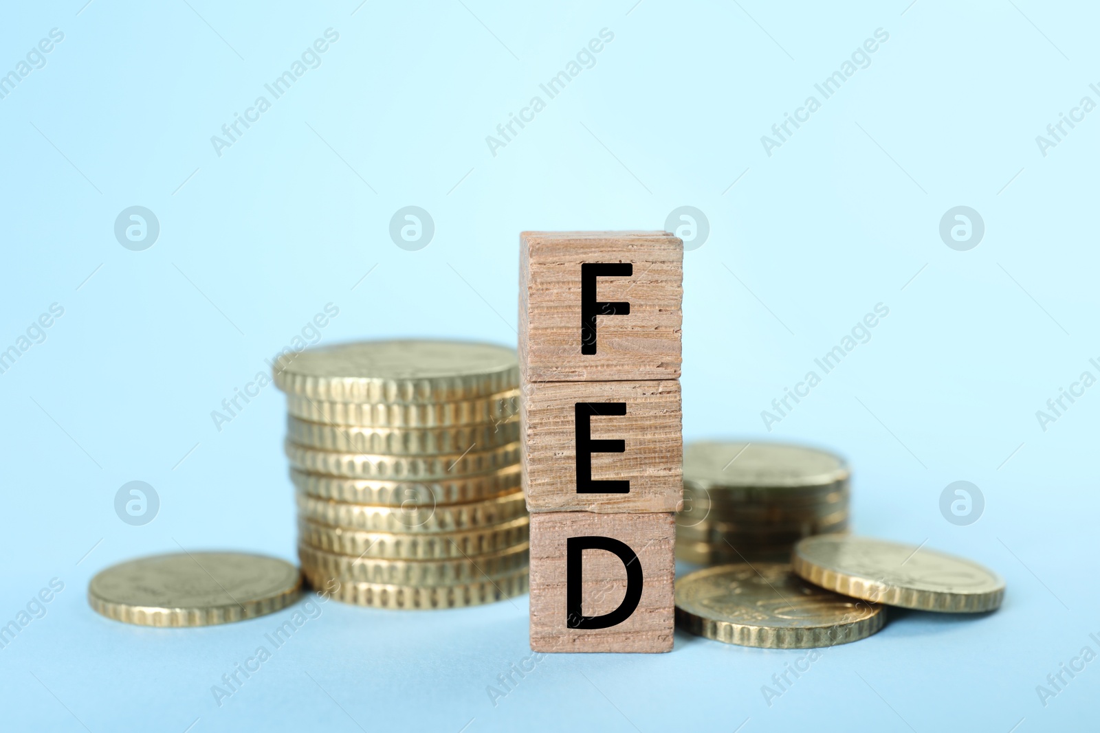 Photo of Stack of wooden cubes with letters Fed (Federal Reserve System) and coins on light blue background, closeup