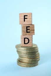 Photo of Stack of wooden cubes with letters Fed (Federal Reserve System) and coins on light blue background, closeup