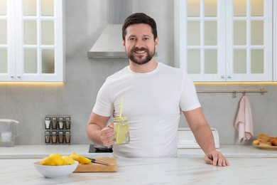 Photo of Happy man holding mason jar of water with lemon in kitchen