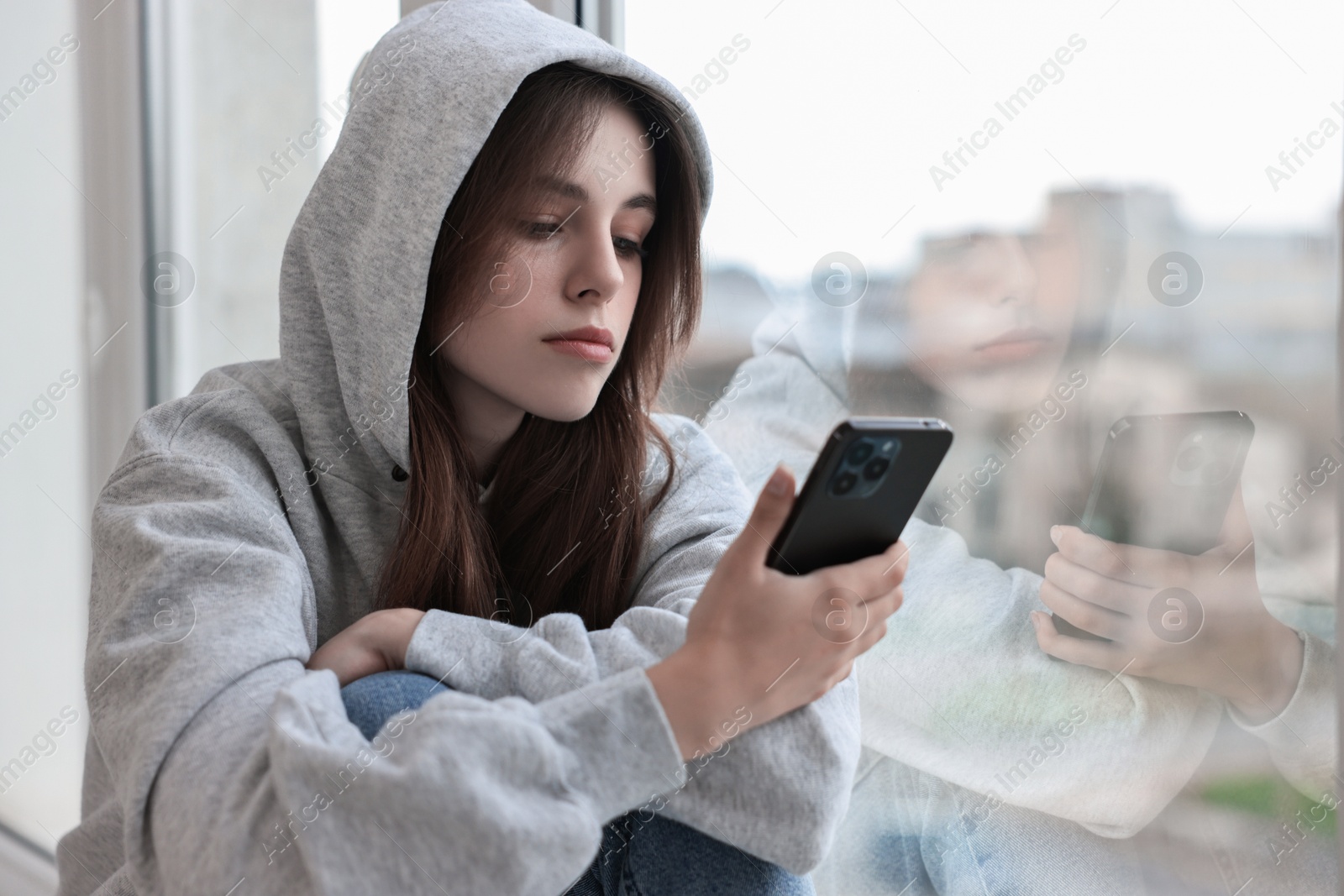 Photo of Loneliness concept. Sad teenage girl using smartphone near window at home