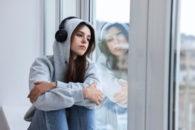 Loneliness concept. Sad teenage girl in headphones listening to music on windowsill at home