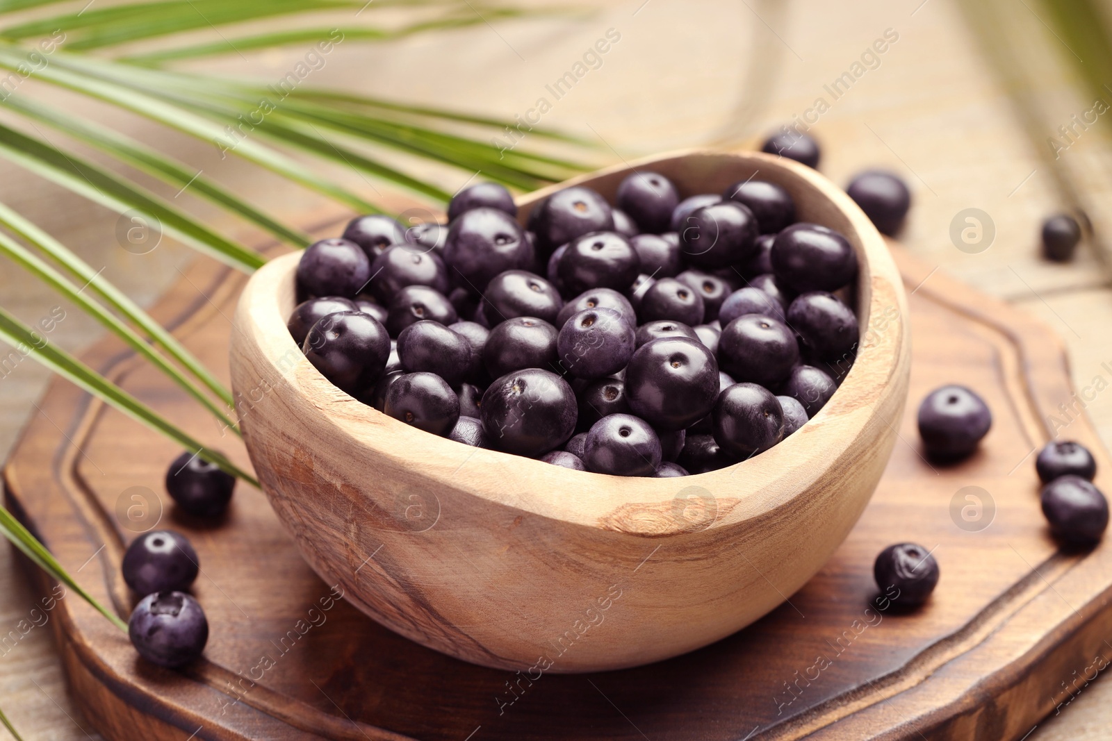 Photo of Ripe acai berries in bowl and palm leaves on wooden table, closeup