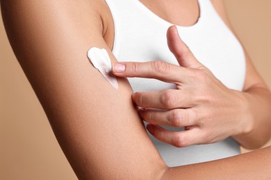 Woman applying cream onto arm against beige background, closeup. Body care