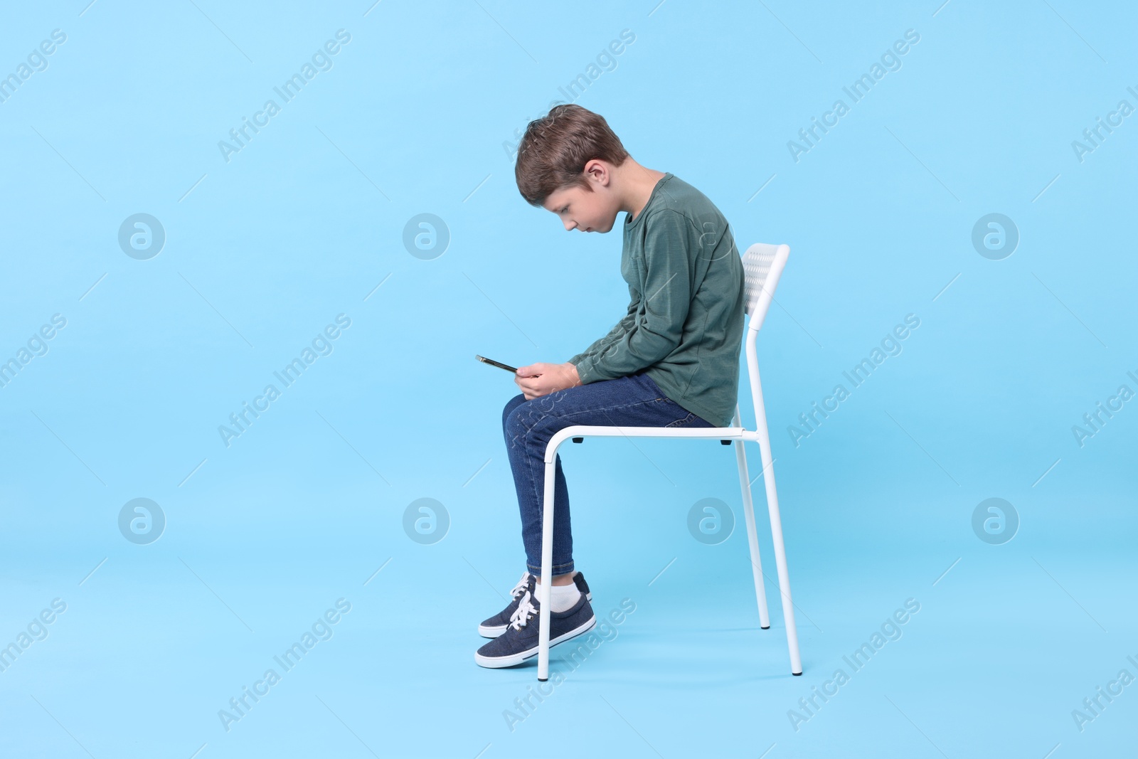 Photo of Boy with incorrect posture and phone sitting on chair against light blue background