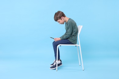 Boy with incorrect posture and phone sitting on chair against light blue background