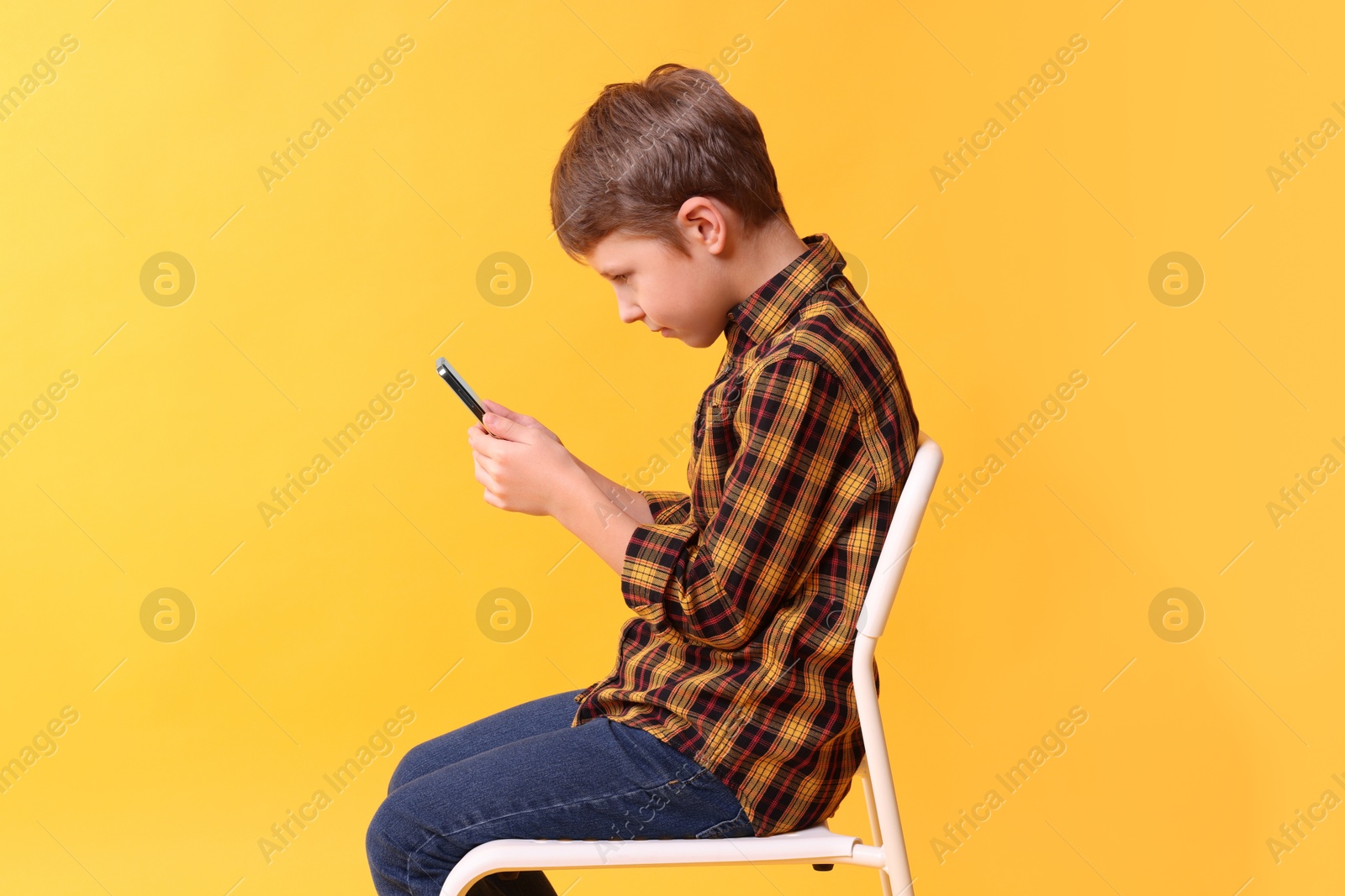 Photo of Boy with incorrect posture and smartphone sitting on chair against yellow background