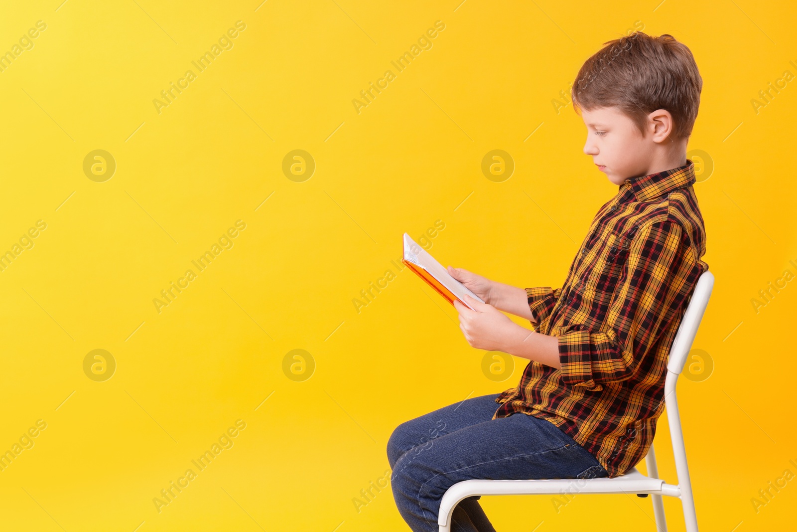 Photo of Boy with correct posture reading book on chair against yellow background