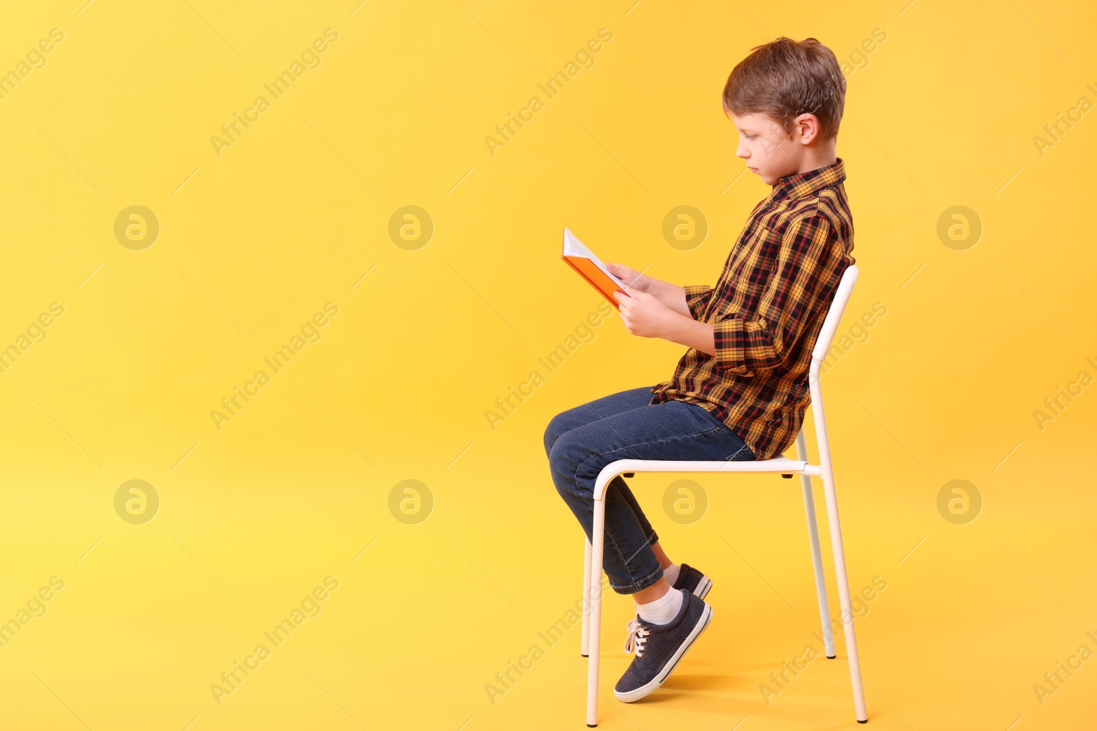 Photo of Boy with correct posture reading book on chair against yellow background