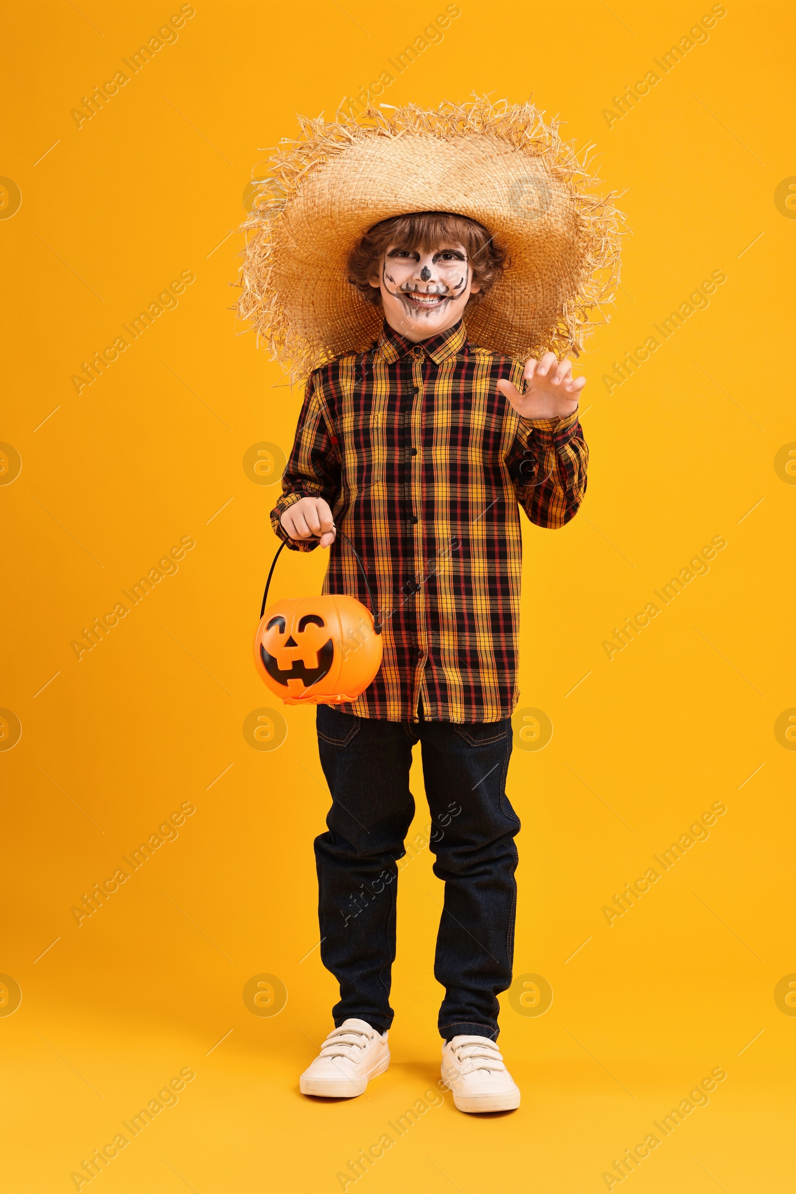 Photo of Funny boy with pumpkin bucket dressed like scarecrow on yellow background. Halloween celebration