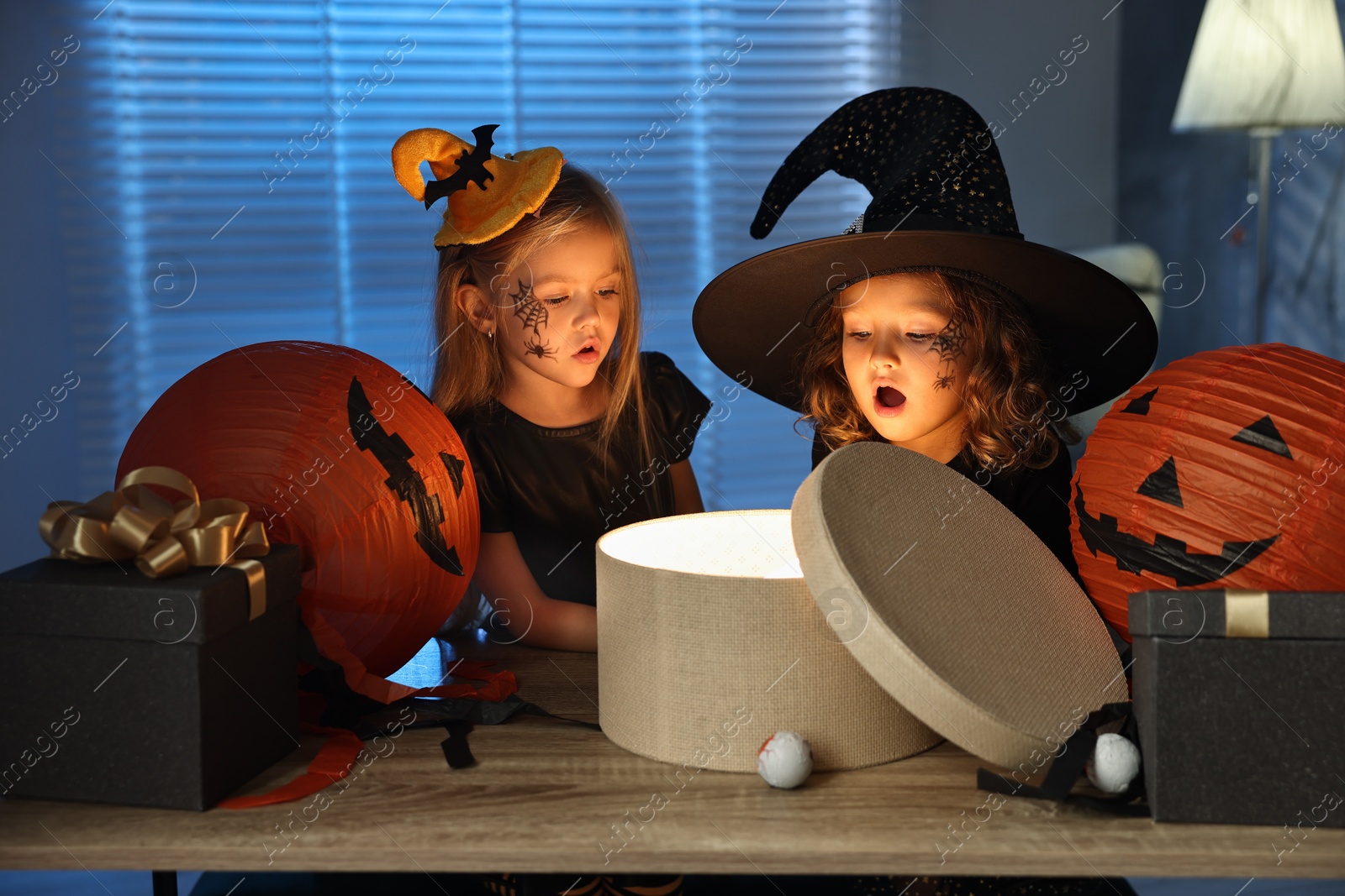 Photo of Surprised children in costumes with festive decor and gift boxes indoors at night. Halloween celebration