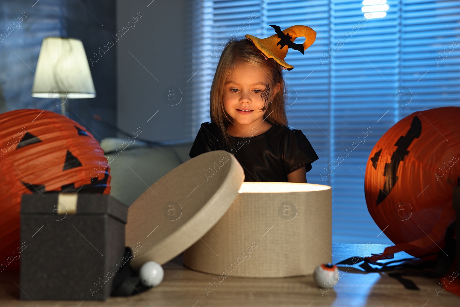 Photo of Cute girl dressed like witch with festive decor and gift boxes indoors at night. Halloween celebration