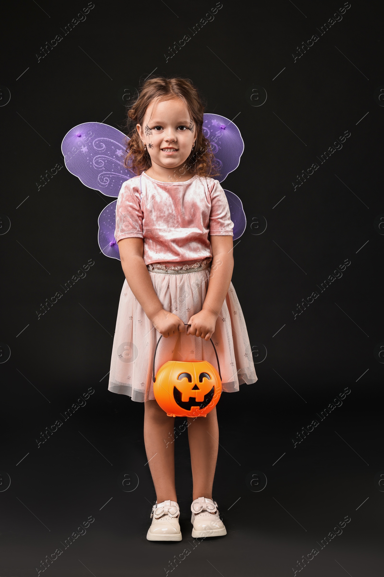 Photo of Cute girl with pumpkin bucket dressed like fairy for Halloween celebration on black background