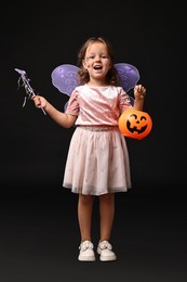 Photo of Cute girl with pumpkin bucket and magic wand dressed like fairy for Halloween celebration on black background