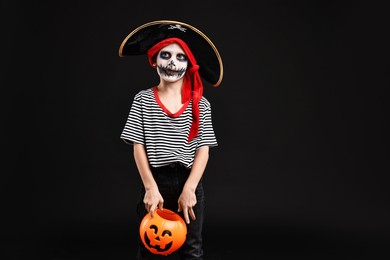 Funny boy with pumpkin bucket dressed like pirate on black background, space for text. Halloween costume