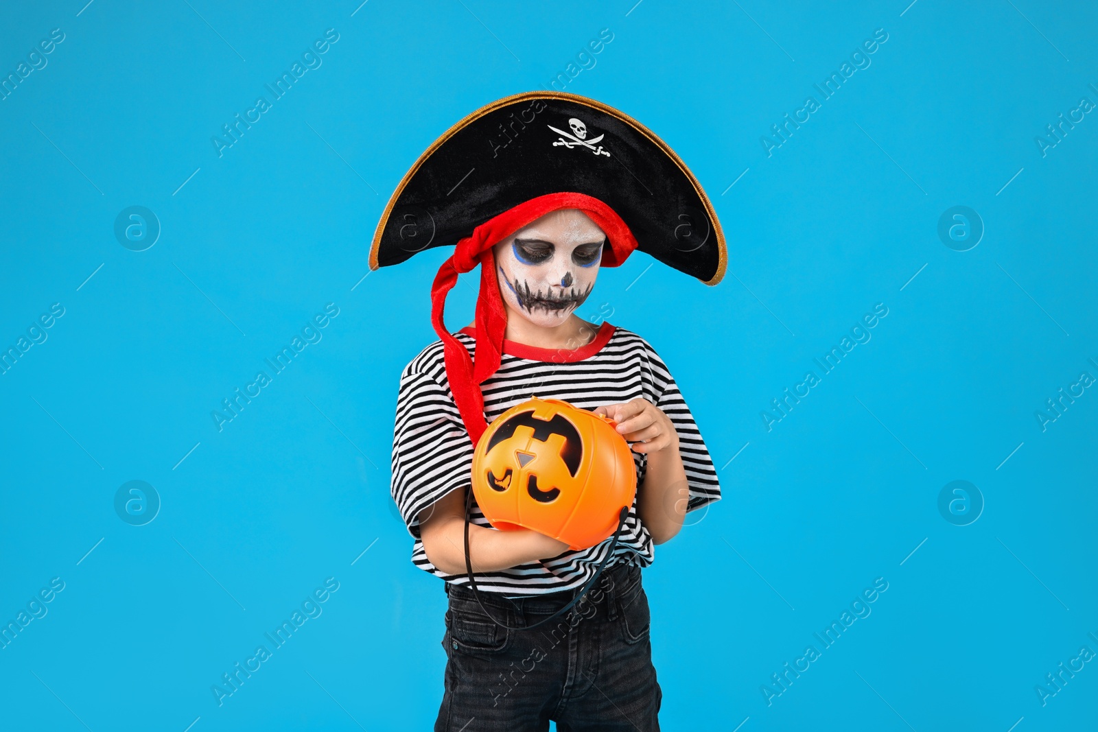 Photo of Funny boy with pumpkin bucket dressed like pirate on light blue background. Halloween costume