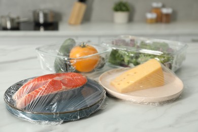 Photo of Different products with plastic food wrap on white marble table in kitchen