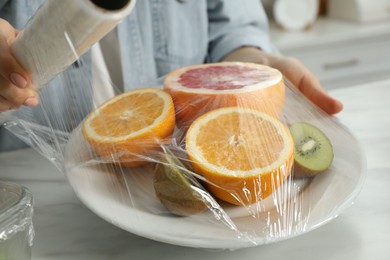 Woman putting plastic food wrap over plate with fresh fruits at white table in kitchen, closeup