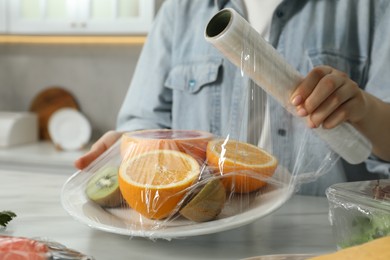 Woman putting plastic food wrap over plate with fresh fruits at white table in kitchen, closeup