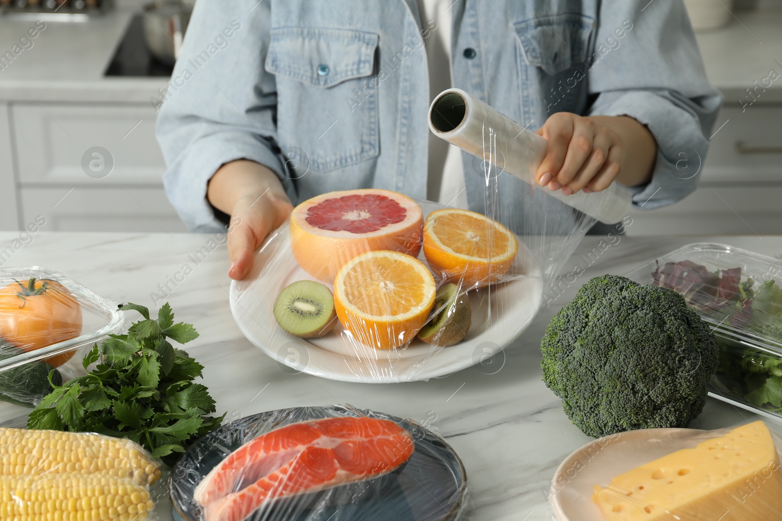 Photo of Woman putting plastic food wrap over plate with fresh fruits at white table in kitchen, closeup