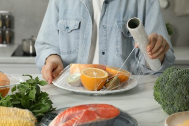 Photo of Woman putting plastic food wrap over plate with fresh fruits at white table in kitchen, closeup