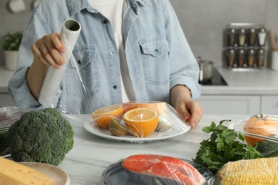 Woman putting plastic food wrap over plate with fresh fruits at white table in kitchen, closeup