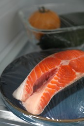 Photo of Plate of salmon with plastic food wrap on shelf in refrigerator, closeup