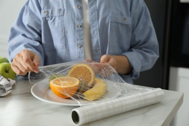 Woman putting plastic food wrap over plate with fruits at countertop in kitchen, closeup