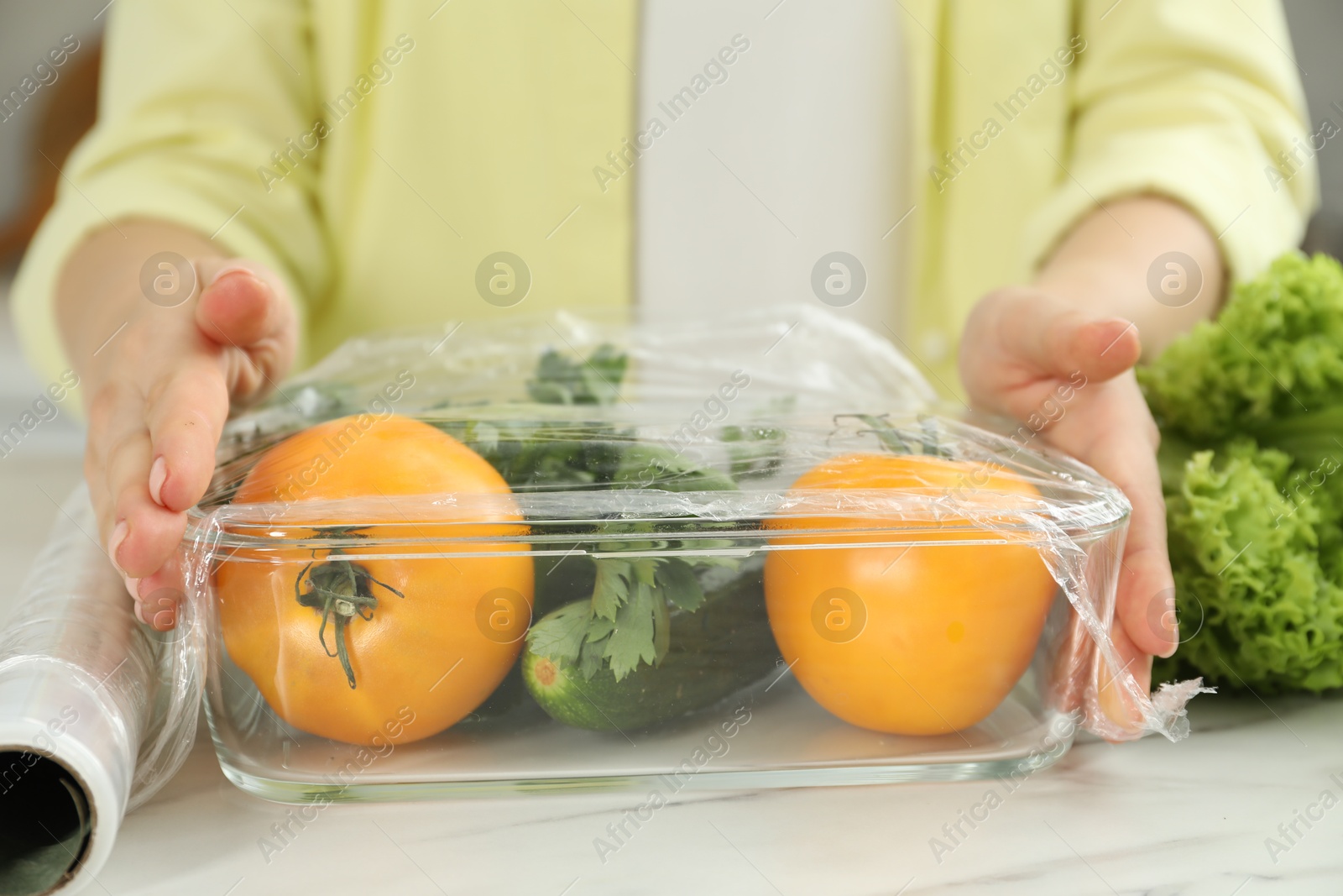 Photo of Woman putting plastic food wrap over glass container with fresh vegetables at white marble table, closeup