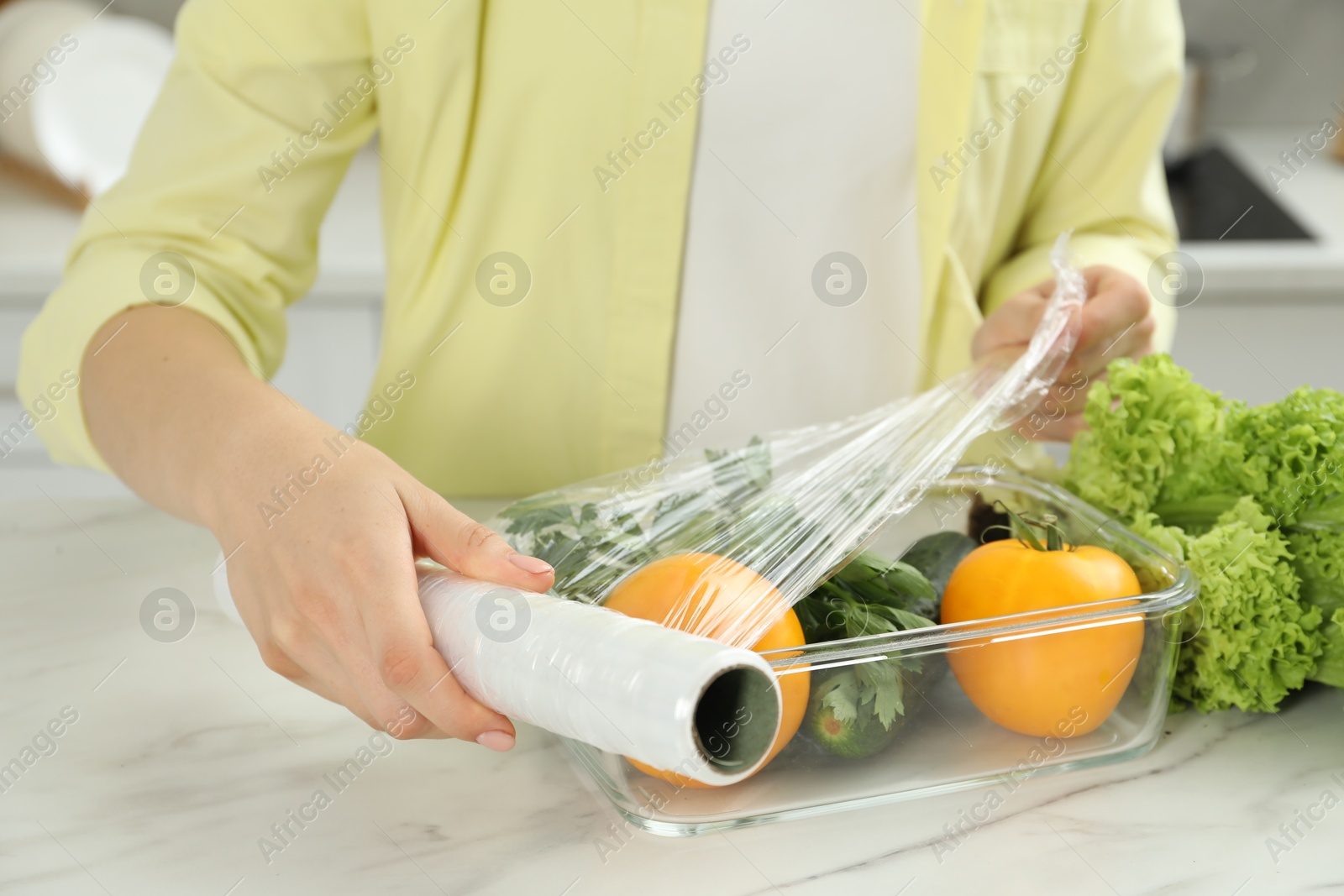 Photo of Woman putting plastic food wrap over glass container with fresh vegetables at white marble table, closeup