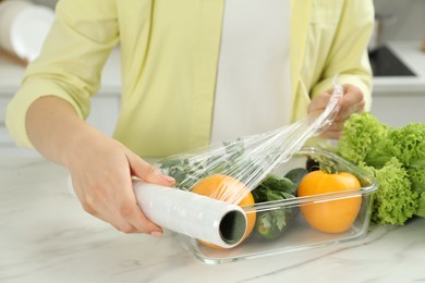 Woman putting plastic food wrap over glass container with fresh vegetables at white marble table, closeup