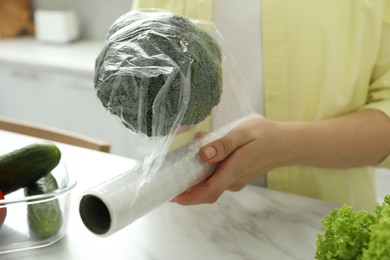 Photo of Woman putting plastic food wrap over broccoli at countertop in kitchen, closeup