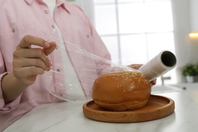 Woman putting plastic food wrap over fresh bun at white marble table in kitchen, closeup