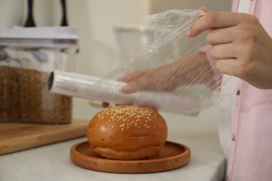 Woman putting plastic food wrap over fresh bun at white table in kitchen, closeup