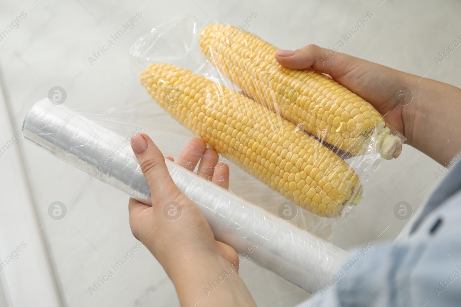 Photo of Woman putting plastic food wrap over corncobs in kitchen, closeup