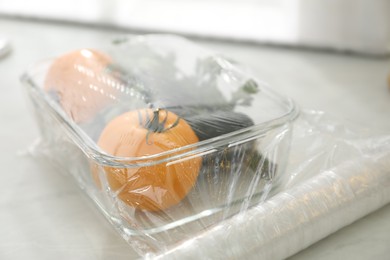 Photo of Glass container of fresh vegetables with plastic food wrap on white table, closeup