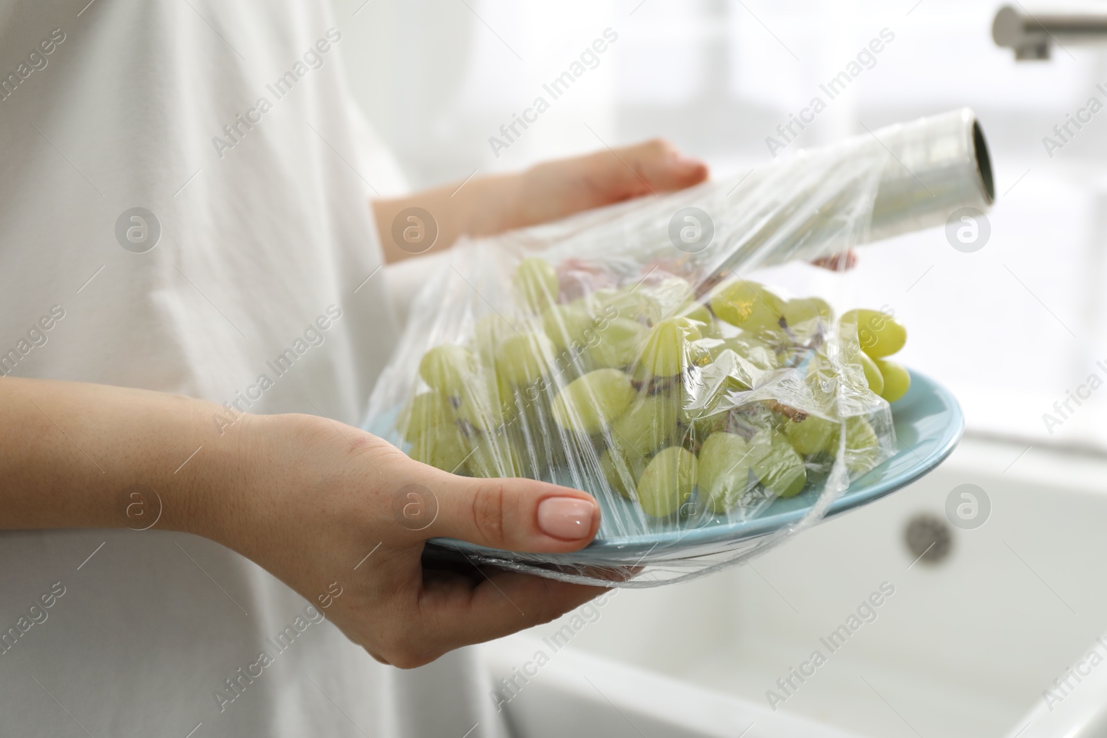 Photo of Woman putting plastic food wrap over plate with grapes in kitchen, closeup