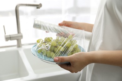 Photo of Woman putting plastic food wrap over plate with grapes in kitchen, closeup