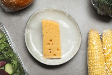 Different products with plastic food wrap on grey table, flat lay