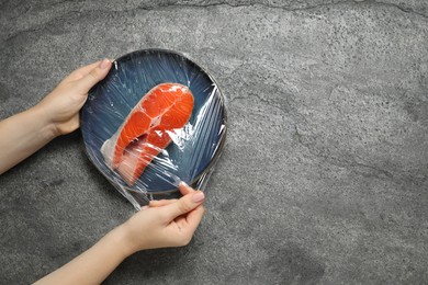 Photo of Woman putting plastic food wrap over plate with salmon at grey table, top view. Space for text