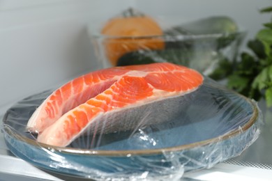 Photo of Plate of salmon with plastic food wrap on shelf in refrigerator, closeup