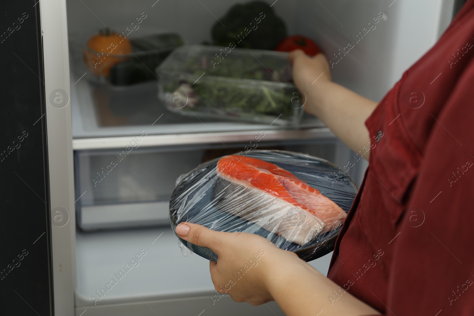 Photo of Woman holding plate of fresh salmon with plastic food wrap near open refrigerator, closeup