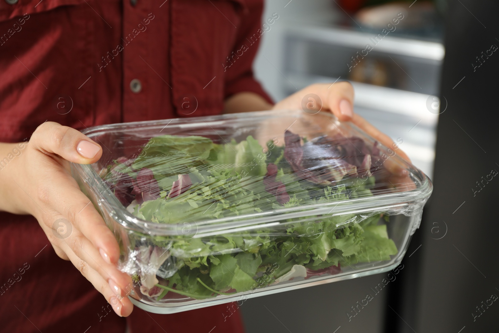 Photo of Woman holding glass container of fresh salad with plastic food wrap in kitchen, closeup