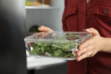 Photo of Woman holding glass container of fresh salad with plastic food wrap in kitchen, closeup