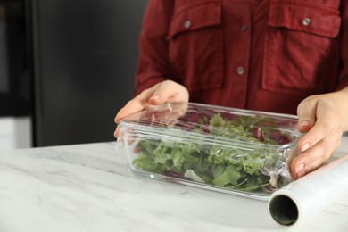 Photo of Woman putting plastic food wrap over glass container with salad at white table in kitchen, closeup